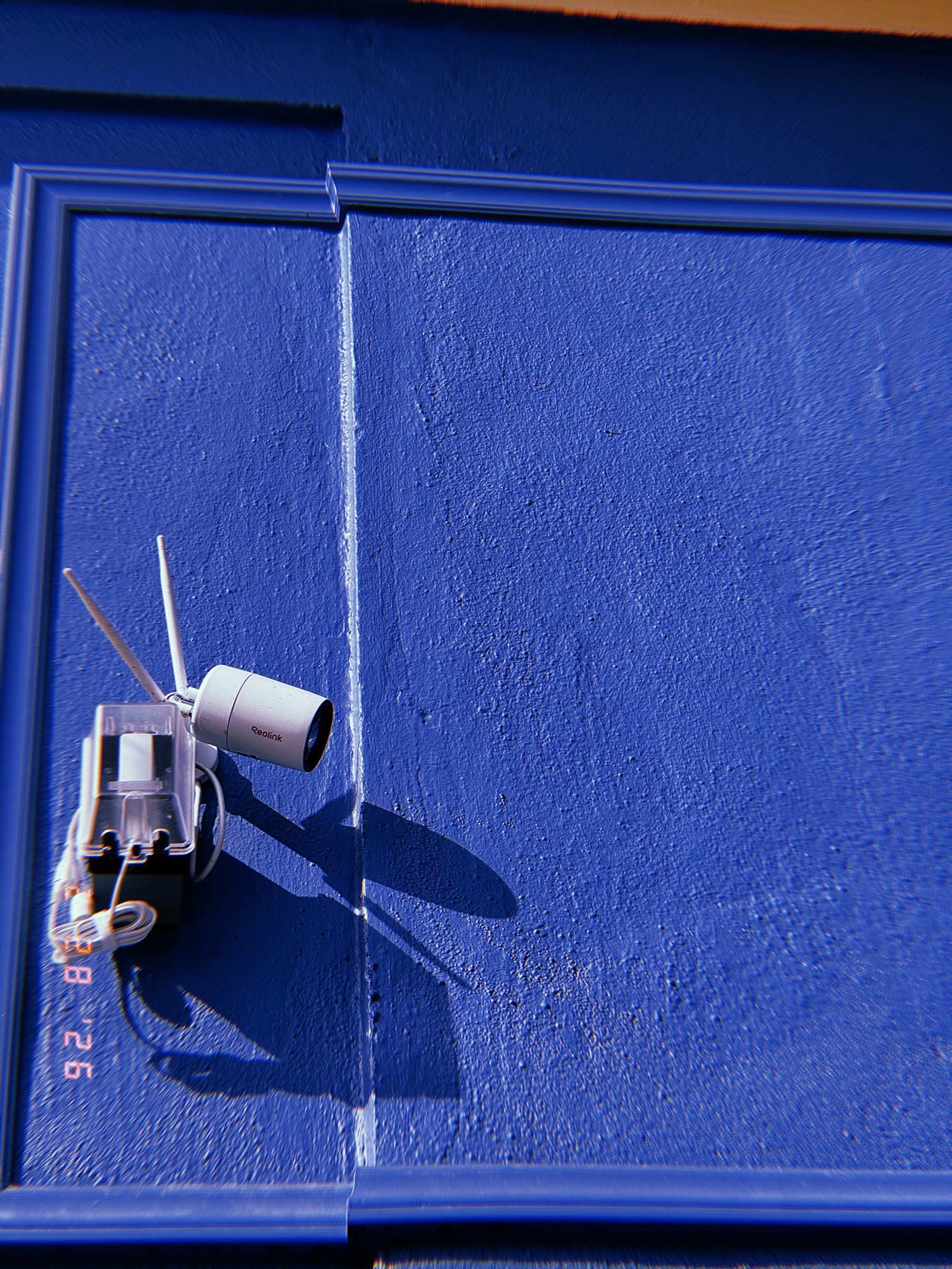 wall-mounted security camera with antenna and transparent junction box casting shadow on blue painted surface