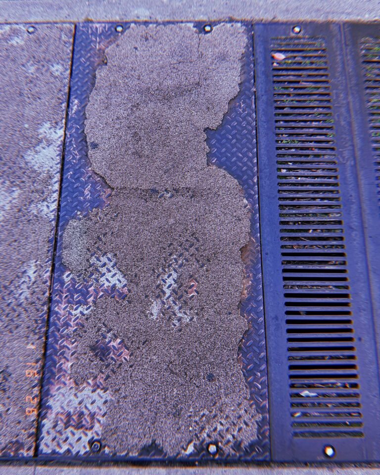 metal sidewalk utility panel partially covered by worn concrete residue next to drainage grate