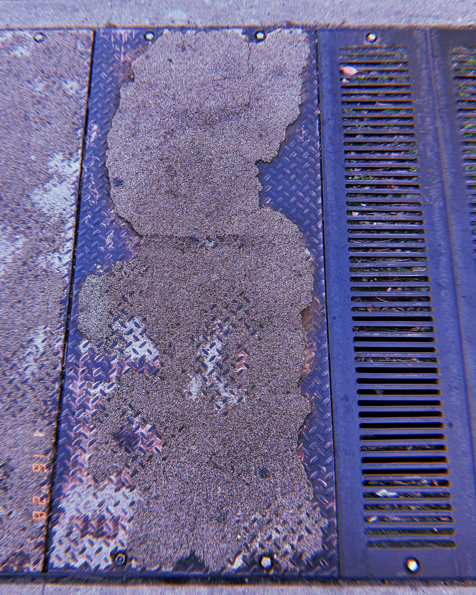 metal sidewalk utility panel partially covered by worn concrete residue next to drainage grate
