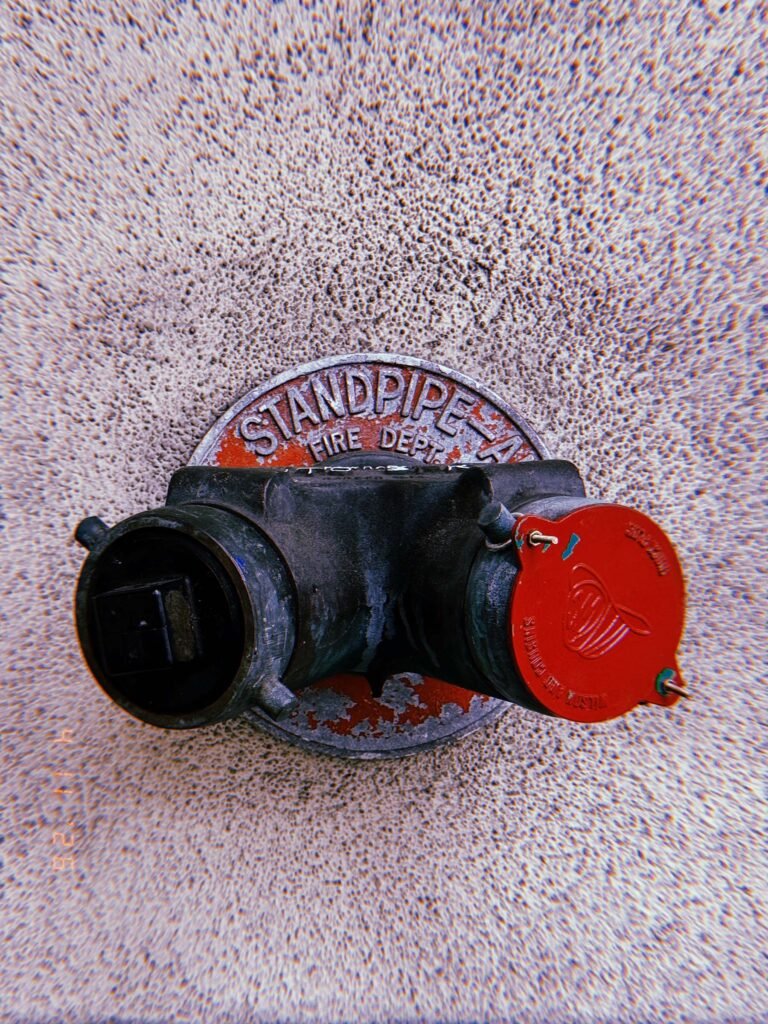 Fire department connection labeled “STANDPIPE” with two hose outlets mounted on a textured wall, one outlet capped in red and the other in black.