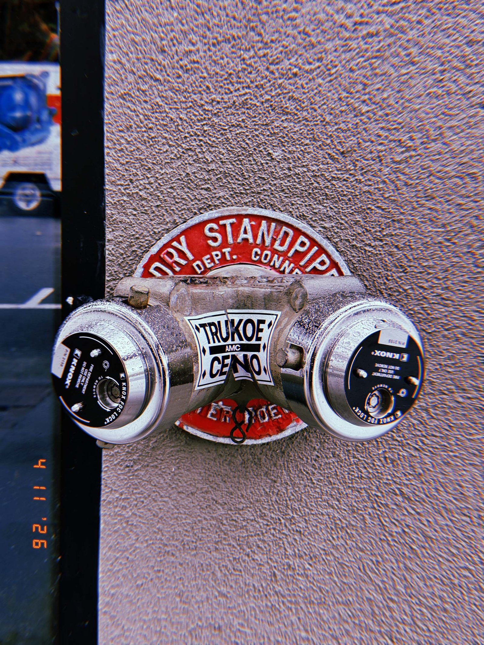 Fire department connection labeled “DRY STANDPIPE” with two capped hose outlets mounted on a textured wall, with a central metal body bearing a sticker.