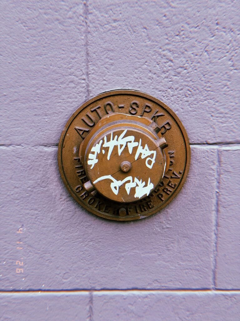 Circular metal fire department connection cover labeled “AUTO SPKR” mounted on a tiled wall, with white graffiti markings on the central cap.