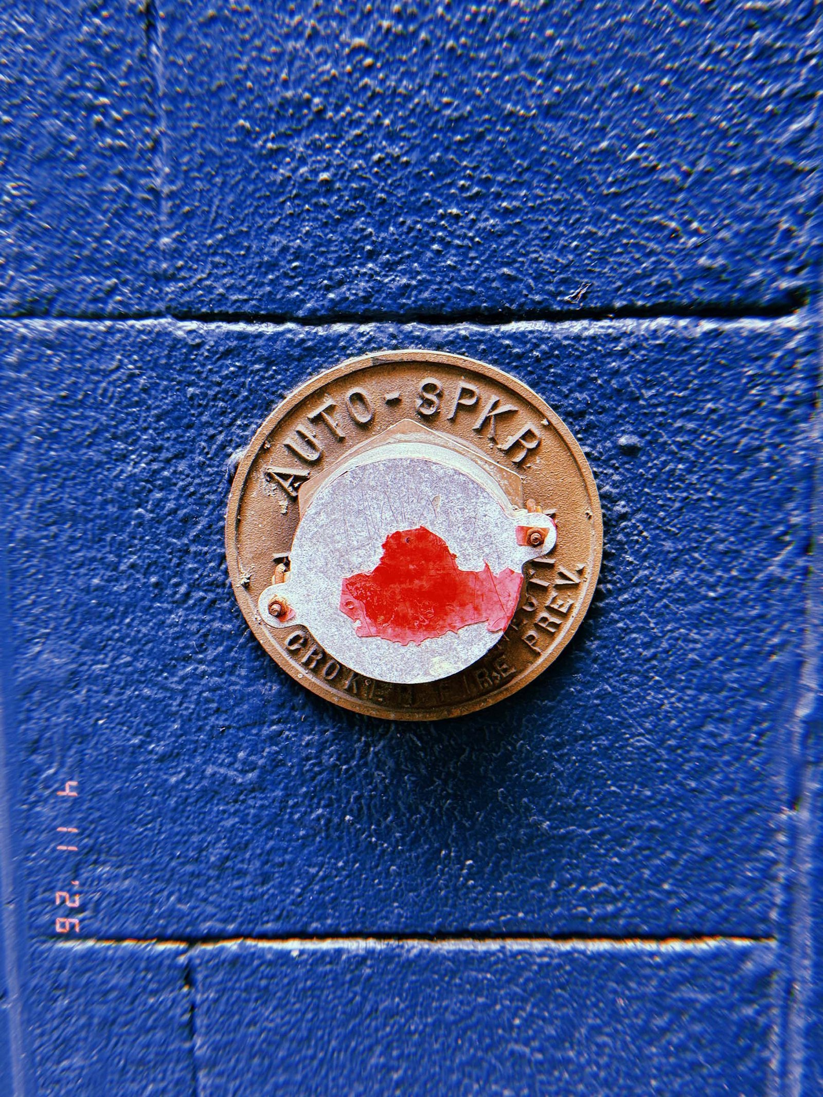Circular metal fire department connection cover labeled “AUTO SPKR” mounted on a blue textured wall, with a central plate showing red paint residue.