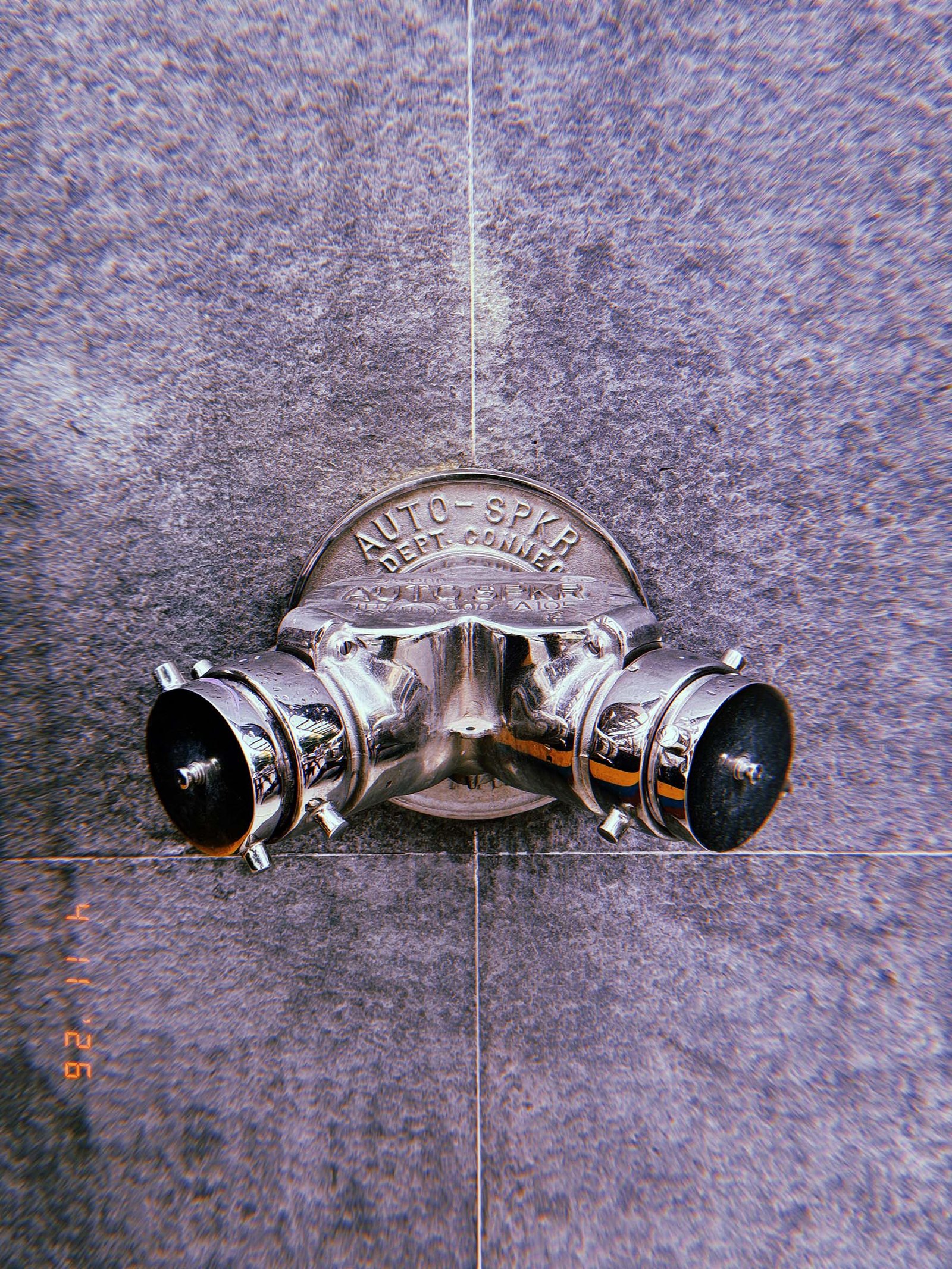Metal fire department connection with two hose outlets mounted on a tiled concrete wall, with a circular plate labeled “AUTO SPKR,” showing polished surface and reflections.
