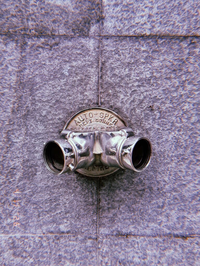 Metal fire department connection with two hose outlets mounted on a tiled concrete wall, with a circular plate labeled “AUTO SPKR,” showing polished surface and reflections.