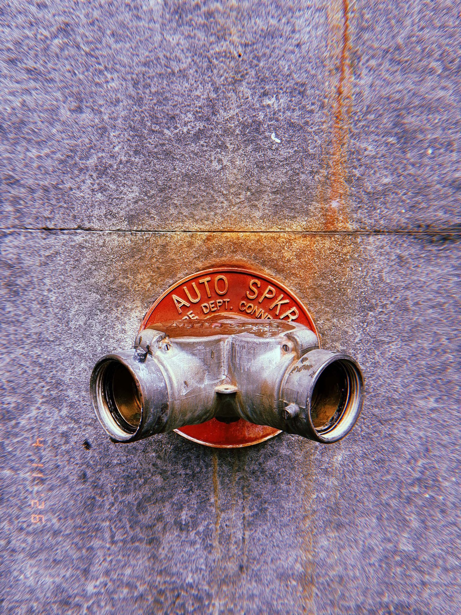 Metal fire department connection with two hose outlets mounted on a concrete wall, set against a red circular plate labeled “AUTO SPKR,” with visible water stains and surface wear.