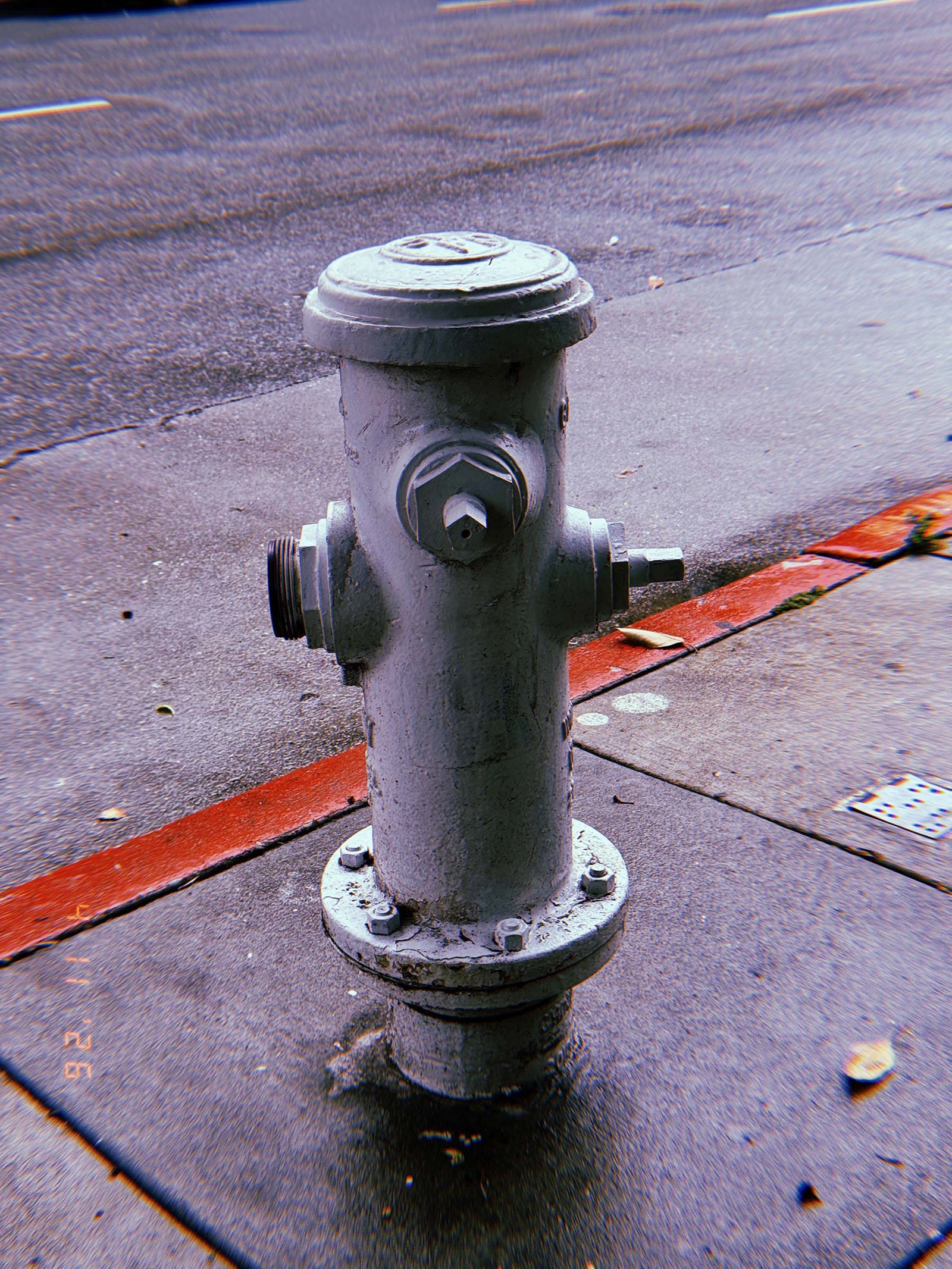 White fire hydrant with exposed threaded outlet and missing caps standing on a sidewalk next to a red-painted curb, with asphalt road in the background.