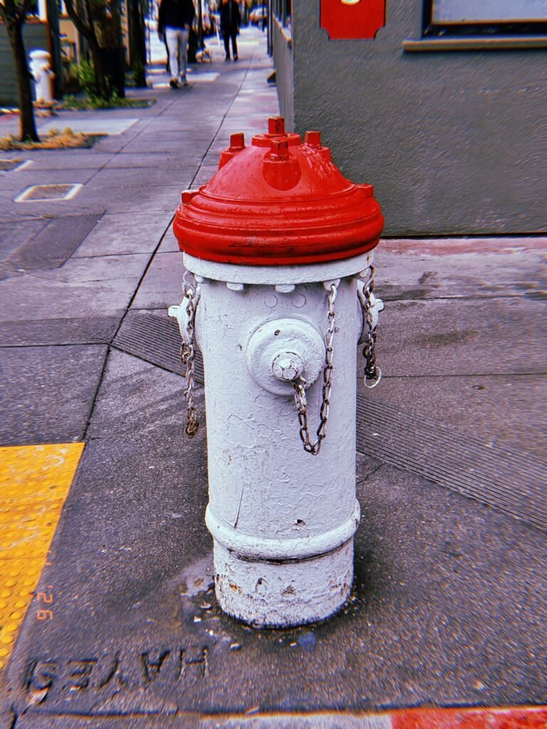White fire hydrant with red cap standing on a sidewalk corner, with chains hanging from the side outlets and a textured pavement surface around it.