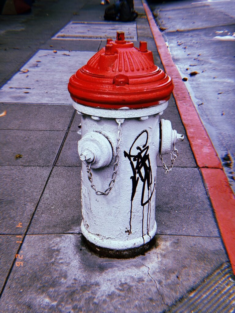 White fire hydrant with red cap standing on a sidewalk next to a red-painted curb, with black graffiti markings and chains attached to the side outlets.