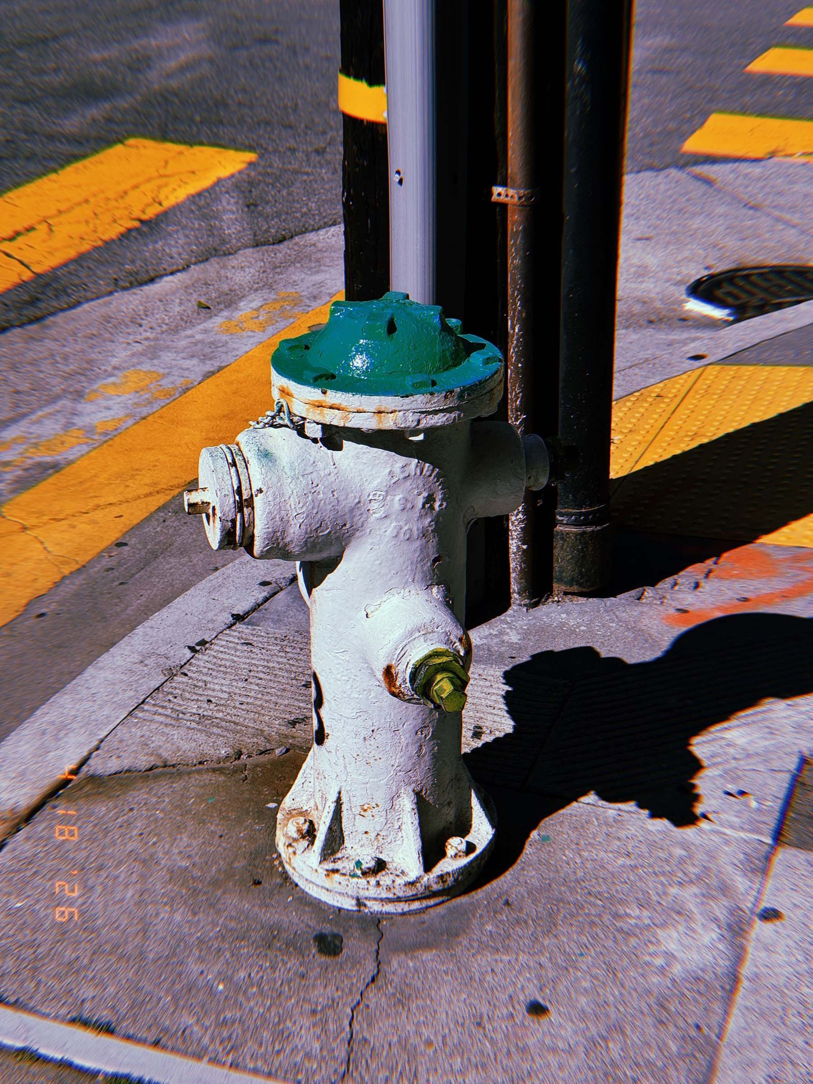 White fire hydrant with green cap mounted on a sidewalk at a street corner next to a metal pole, with yellow curb markings and asphalt road visible.