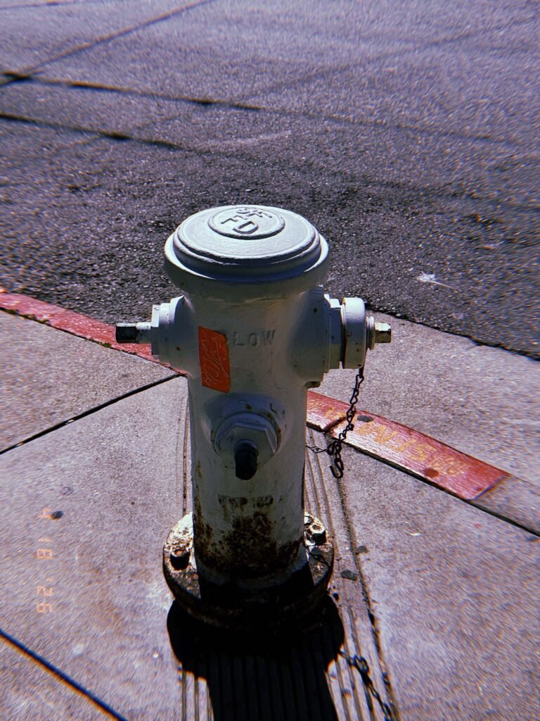 White fire hydrant labeled “LOW” standing on a sidewalk at a street corner, with asphalt road and curb in the background and a chain attached to one outlet.