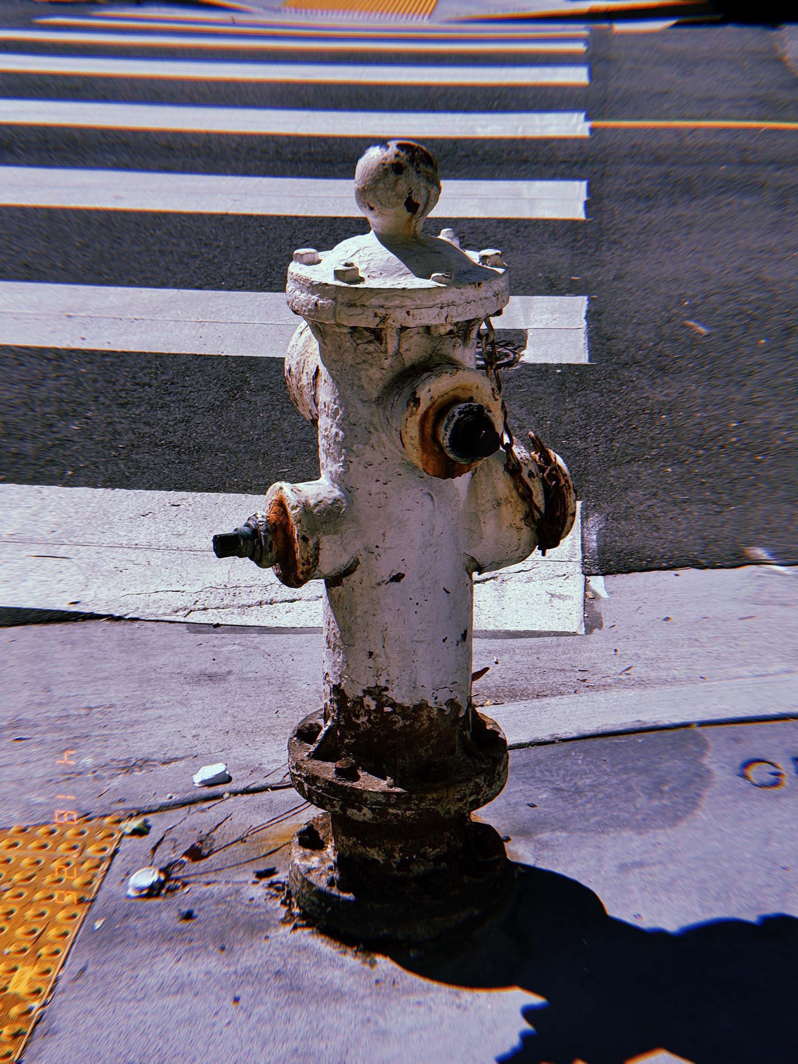 White fire hydrant with rusted fittings standing on a sidewalk next to a crosswalk, with asphalt road and painted pedestrian lines in the background.