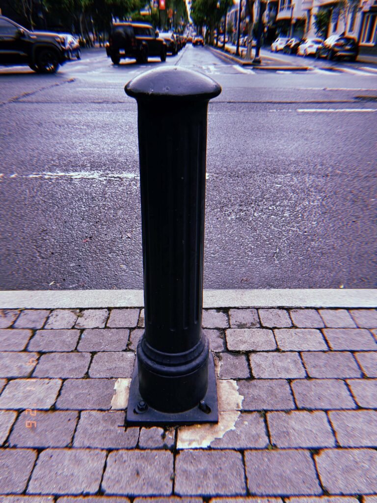 Black metal bollard with rounded top mounted on a cobblestone sidewalk edge, with asphalt road and parked cars in the background.