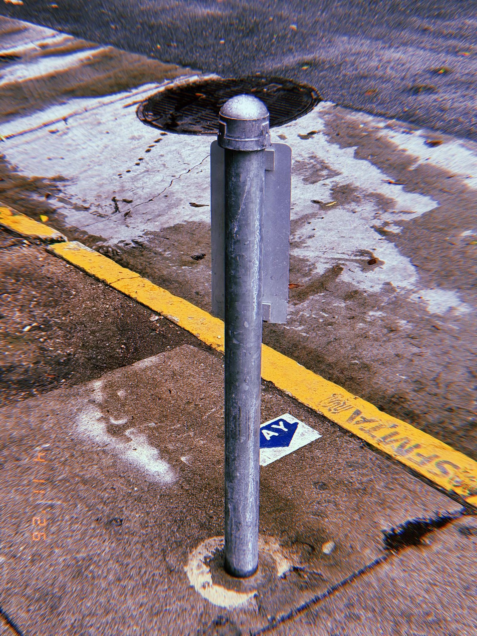 Metal bollard with rounded cap and attached rectangular plate installed on a sidewalk near a yellow-painted curb and street drain.