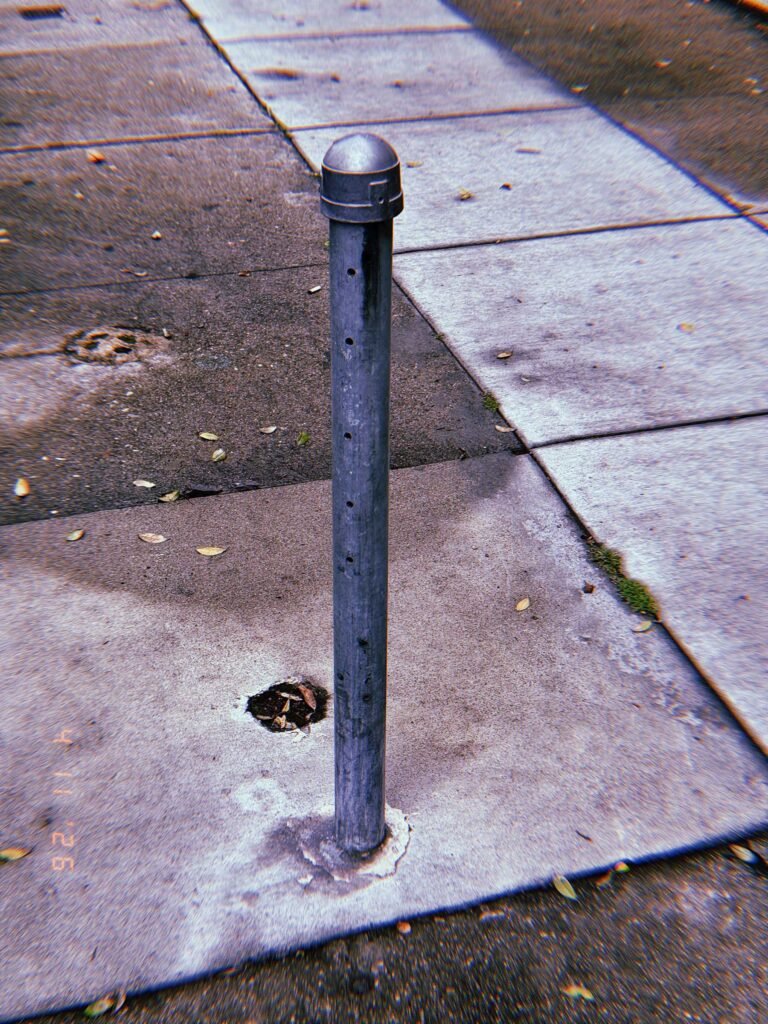 Slim metal bollard with rounded cap installed in a sidewalk, with small perforations along its surface and concrete pavement surrounding it.
