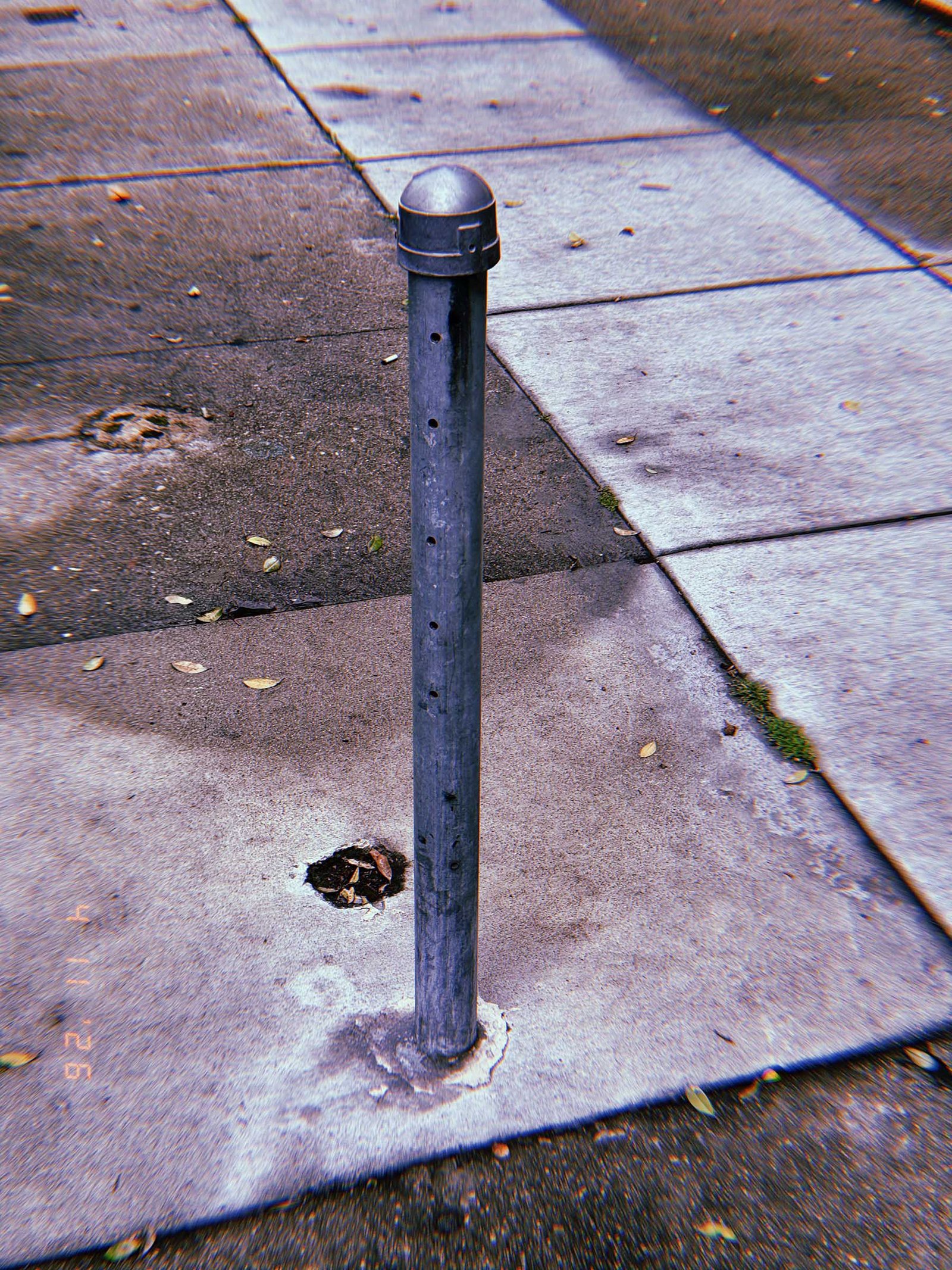 Slim metal bollard with rounded cap installed in a sidewalk, with small perforations along its surface and concrete pavement surrounding it.