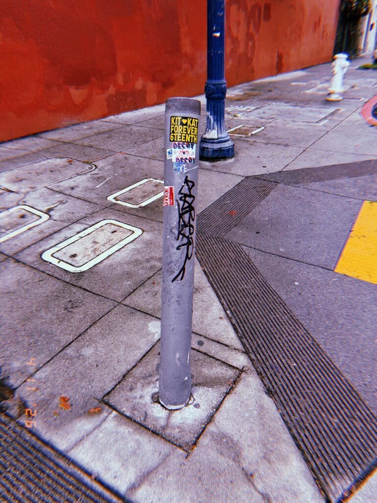 Cylindrical metal bollard with stickers and graffiti installed on a sidewalk corner, with textured pavement and street markings nearby.