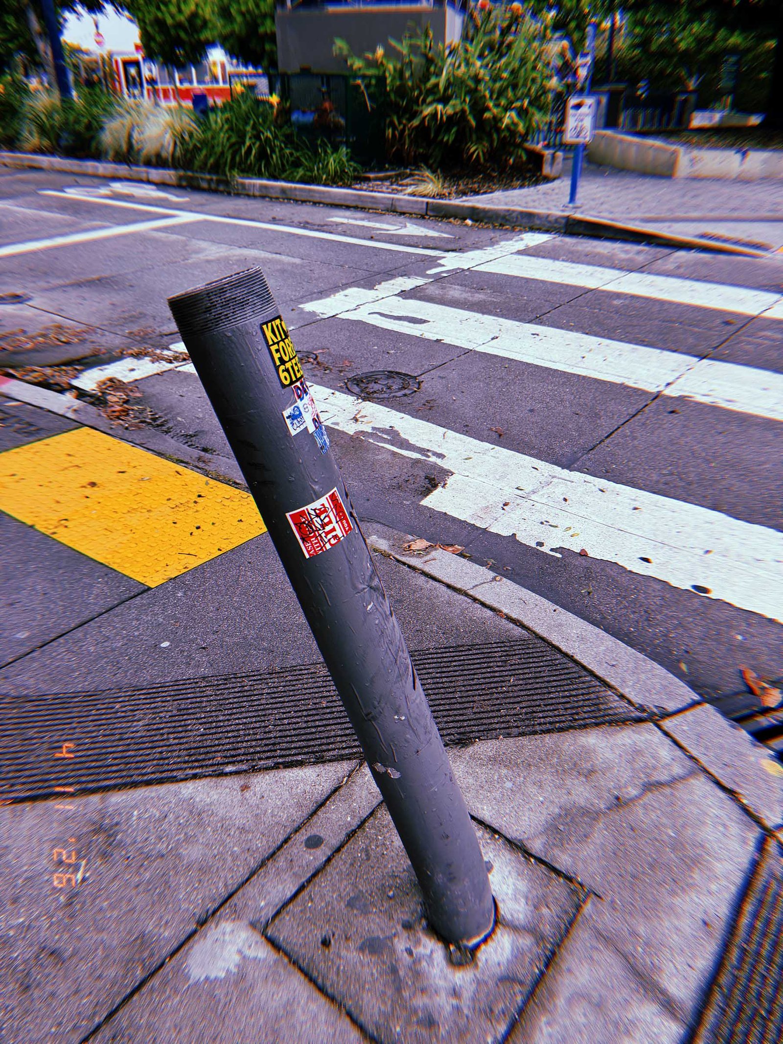 Bent cylindrical metal bollard leaning at an angle on a sidewalk corner near a crosswalk, with stickers attached to its surface.