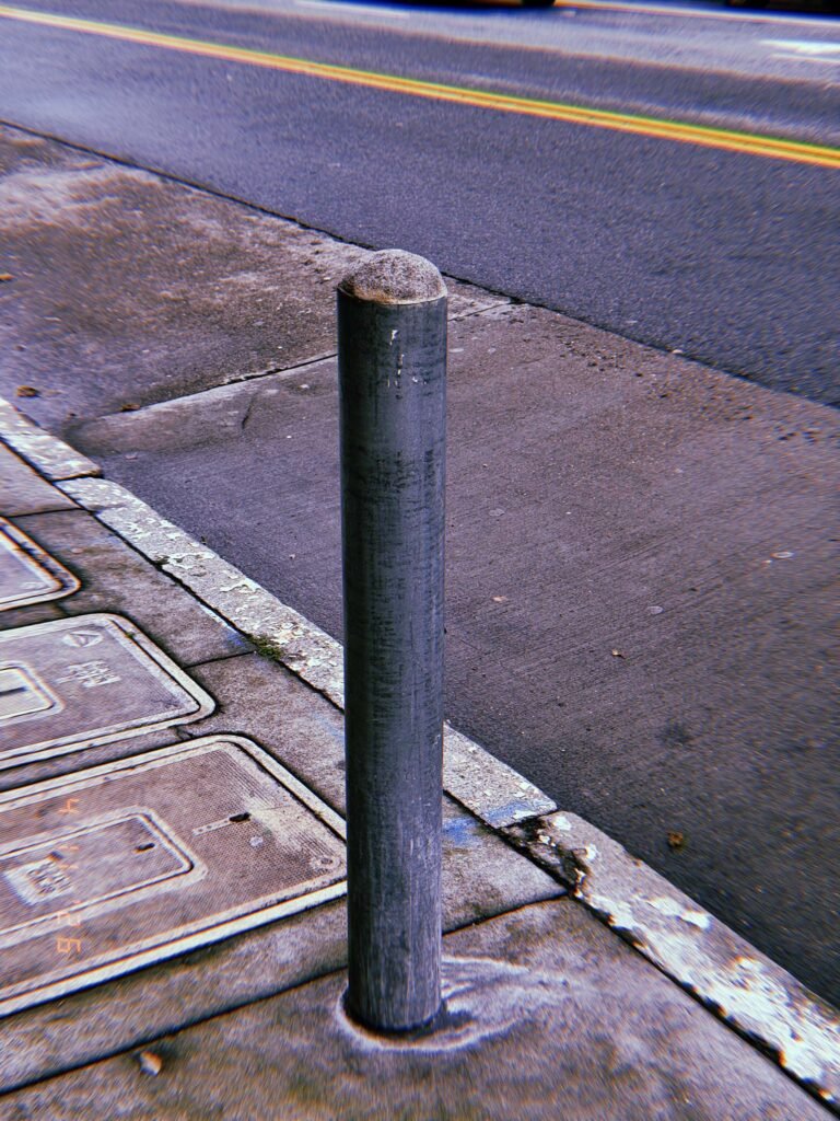 Single cylindrical metal bollard installed on a sidewalk edge next to a curb, with asphalt road and utility covers nearby.