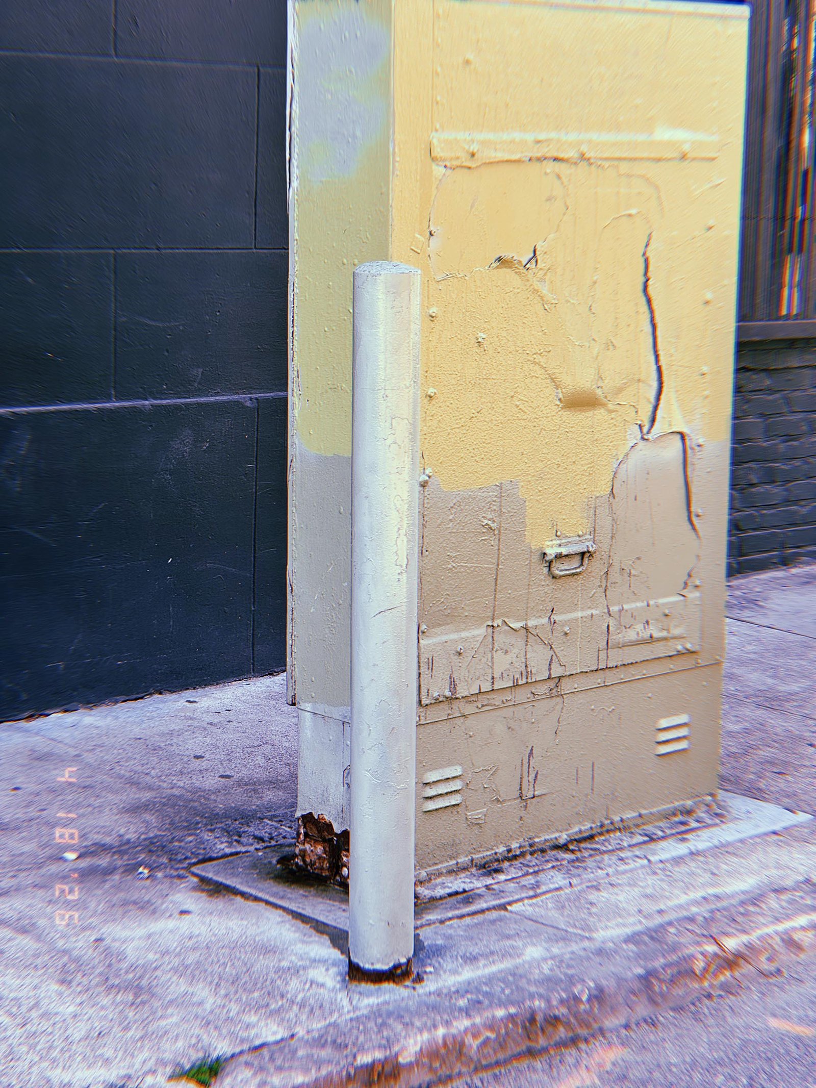 Metal utility cabinet with peeling paint mounted on a sidewalk, protected by a cylindrical metal bollard positioned in front of it.