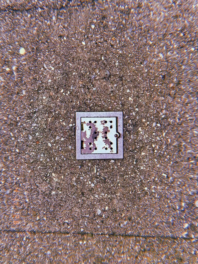 Square perforated metal surface drain embedded in exposed aggregate concrete sidewalk with circular discoloration surrounding the inlet frame.