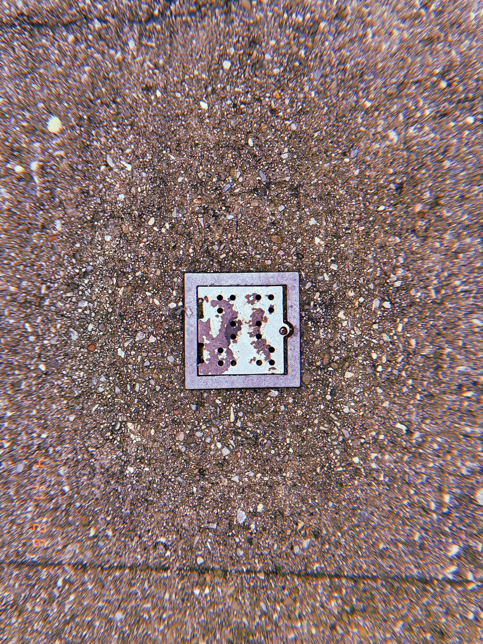 Square perforated metal surface drain embedded in exposed aggregate concrete sidewalk with circular discoloration surrounding the inlet frame.