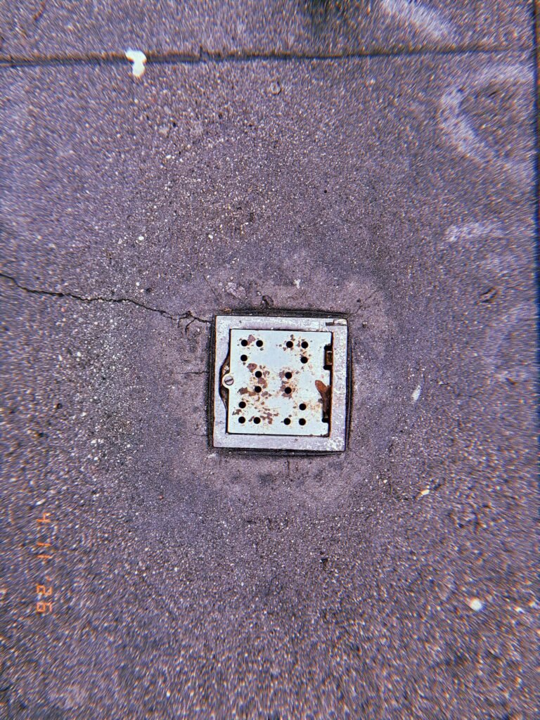 Square perforated metal surface drain embedded in textured concrete sidewalk with surrounding pavement cracks and rust marks.