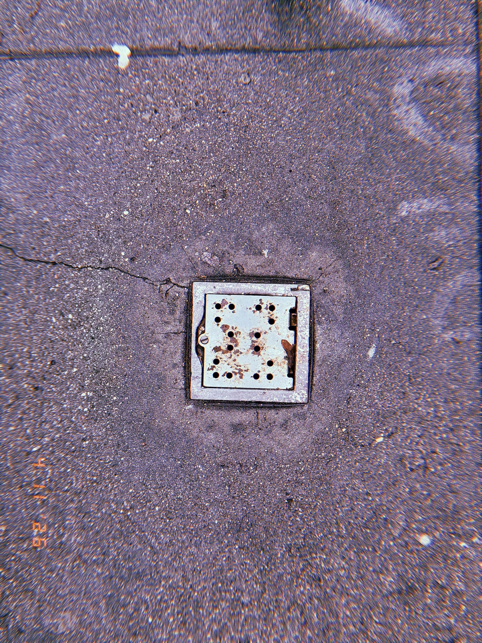 Square perforated metal surface drain embedded in textured concrete sidewalk with surrounding pavement cracks and rust marks.