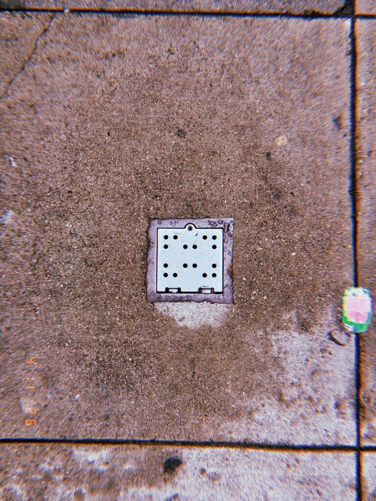 Square perforated metal surface drain embedded in concrete sidewalk with surrounding discoloration and small painted curb marking nearby.