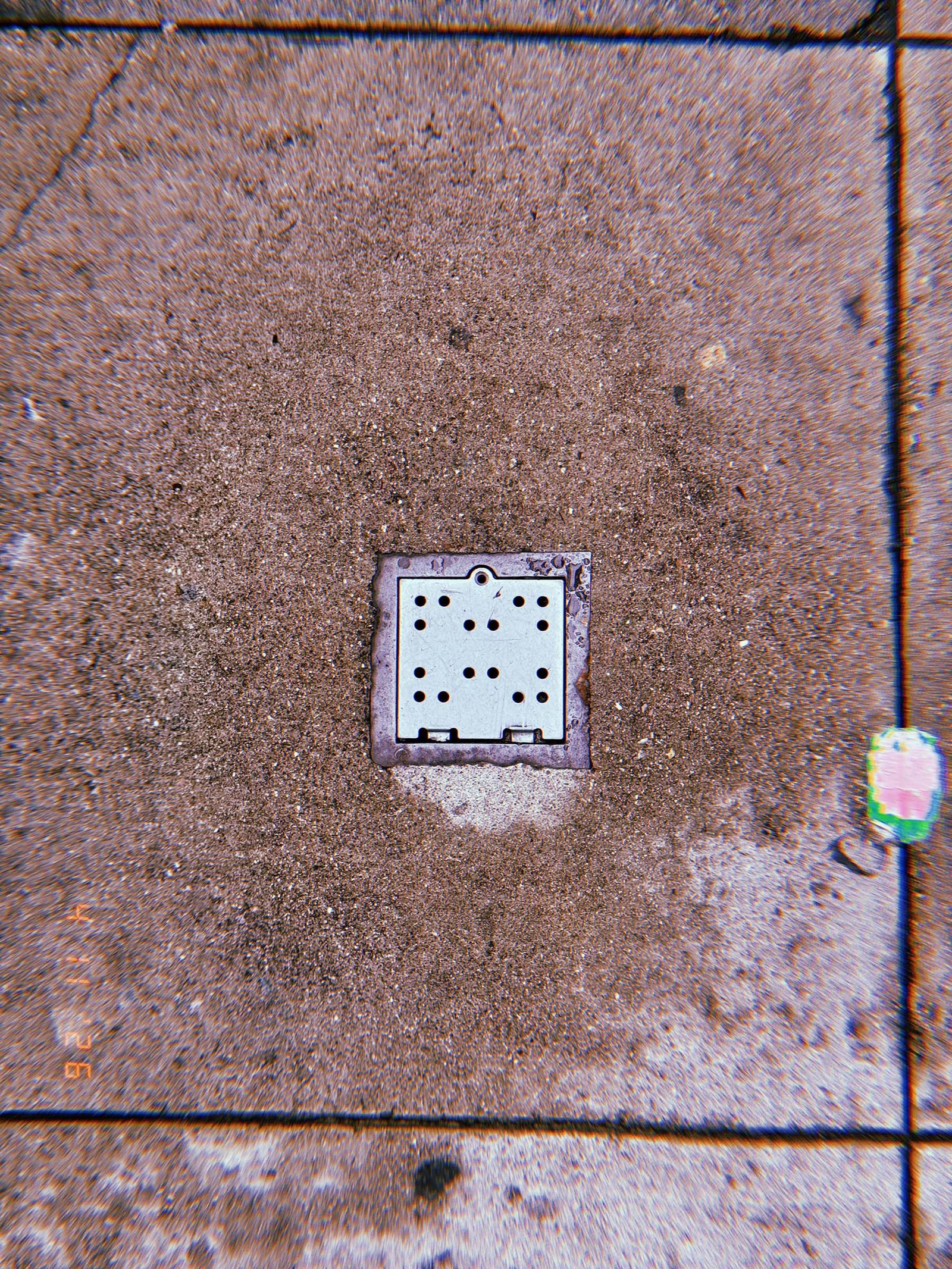 Square perforated metal surface drain embedded in concrete sidewalk with surrounding discoloration and small painted curb marking nearby.