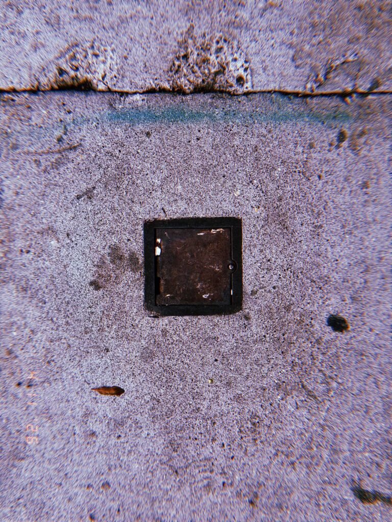 Square metal utility drain cover embedded in concrete sidewalk with dark frame and surrounding surface stains.