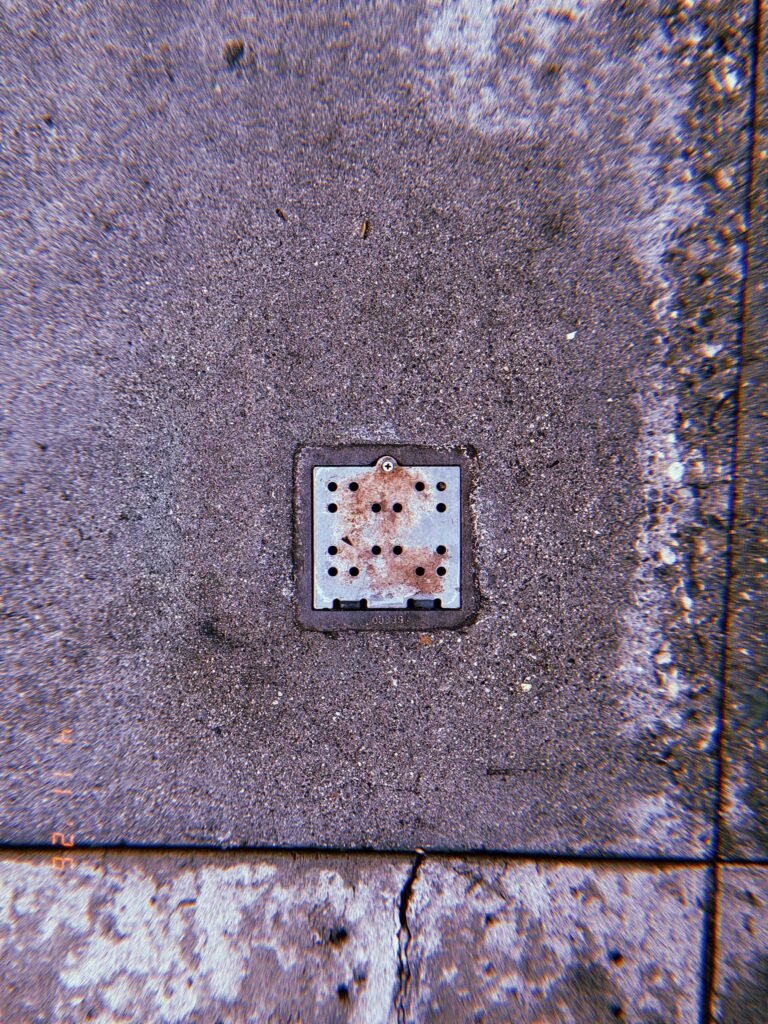 Square perforated metal drain cover embedded in concrete sidewalk with surrounding pavement seams and surface discoloration.