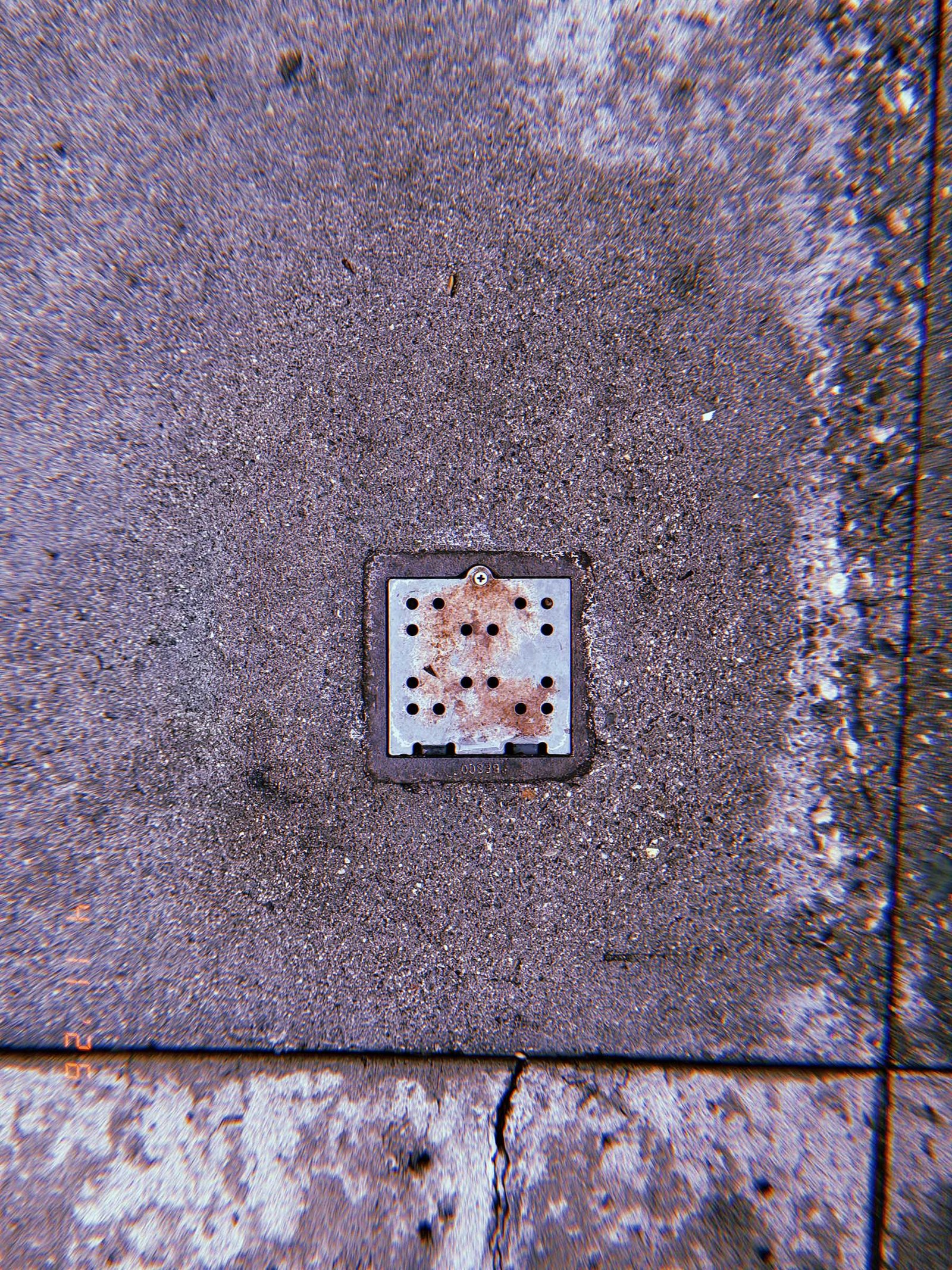 Square perforated metal drain cover embedded in concrete sidewalk with surrounding pavement seams and surface discoloration.