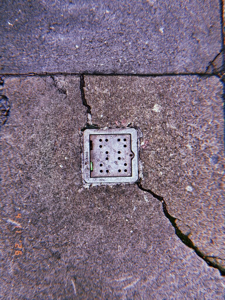 Square perforated surface drain embedded in cracked concrete sidewalk with intersecting pavement fractures surrounding the metal frame.