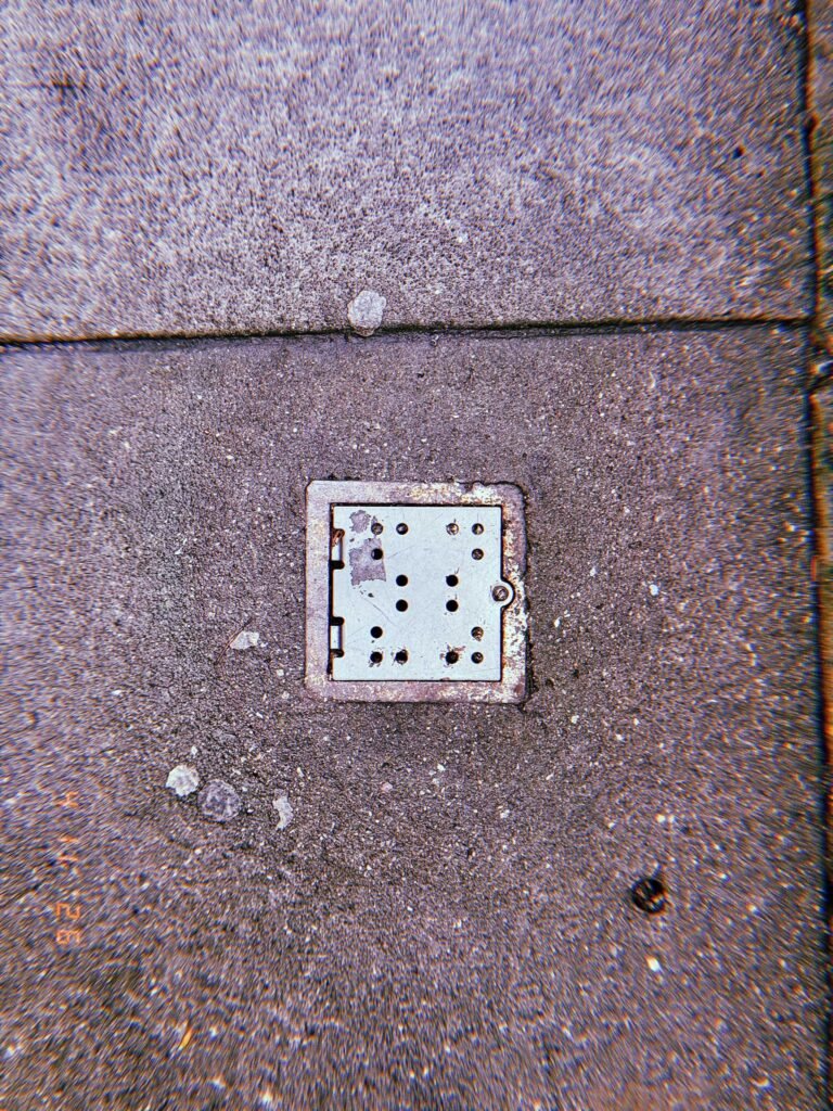 Small square metal surface drain embedded in rough concrete sidewalk with scattered surface marks and pavement seams.