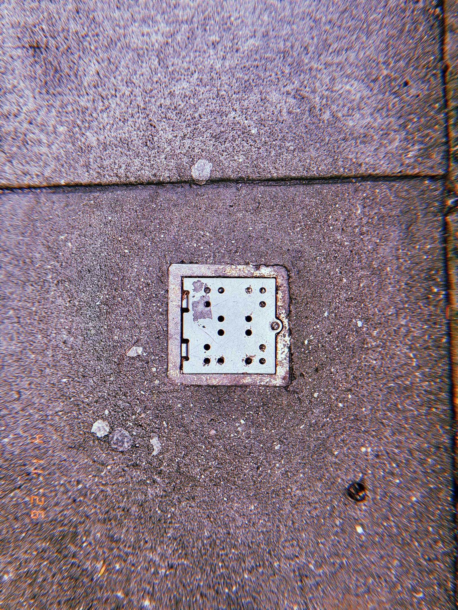 Small square metal surface drain embedded in rough concrete sidewalk with scattered surface marks and pavement seams.
