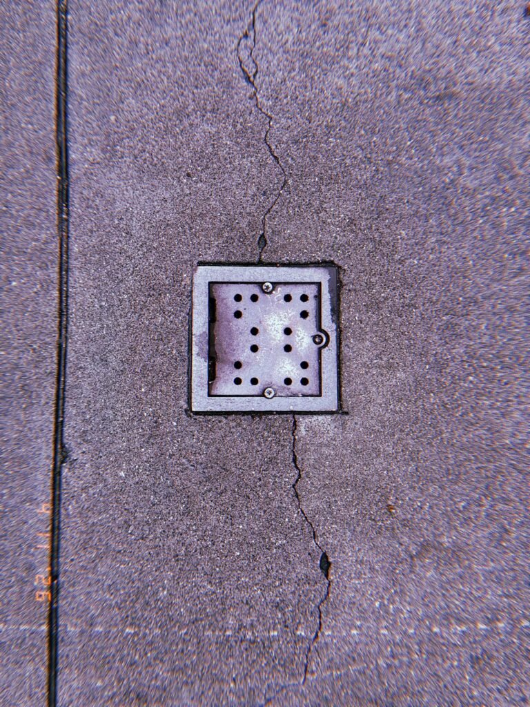 Square perforated metal surface drain embedded in concrete sidewalk, intersected by a vertical pavement crack.