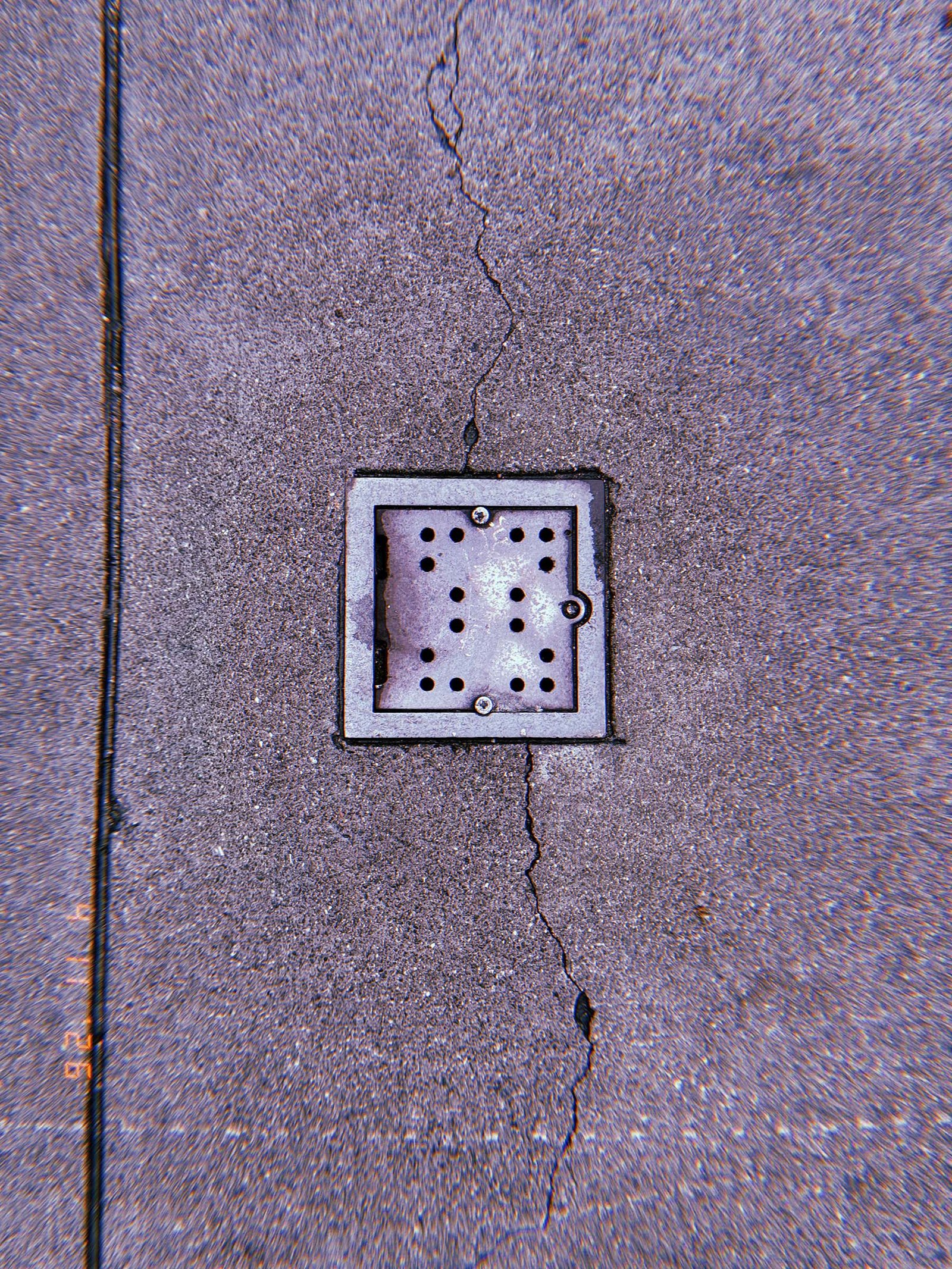 Square perforated metal surface drain embedded in concrete sidewalk, intersected by a vertical pavement crack.