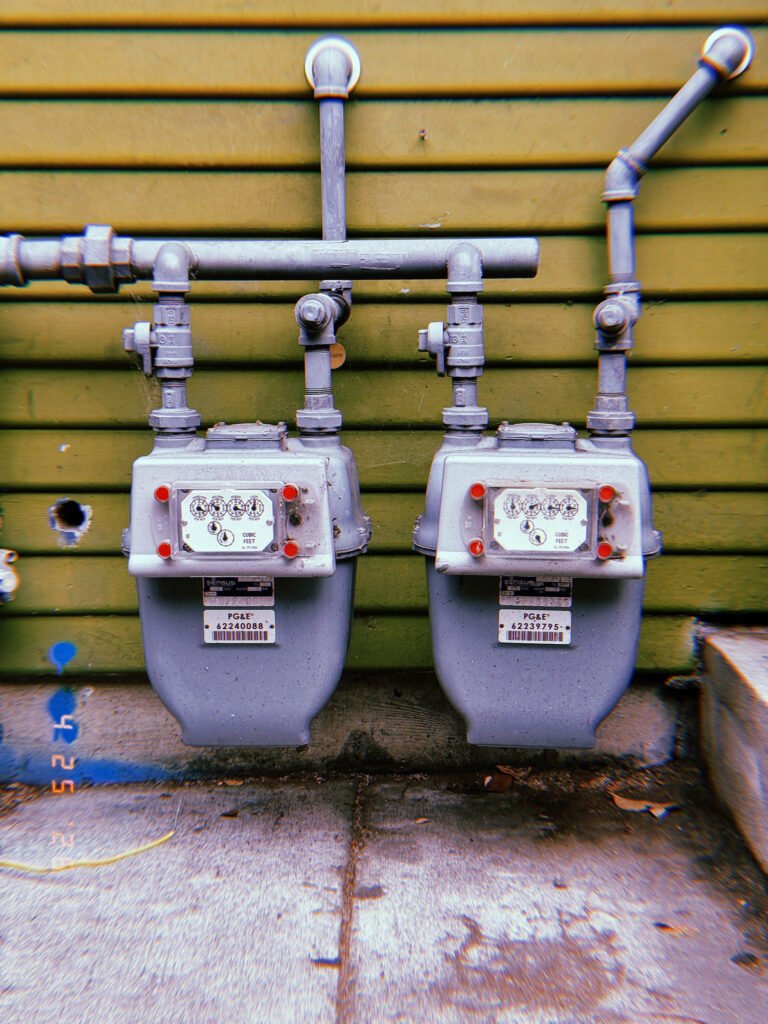 Two residential gas meters mounted side by side beneath exposed utility pipes on a green wooden exterior wall.