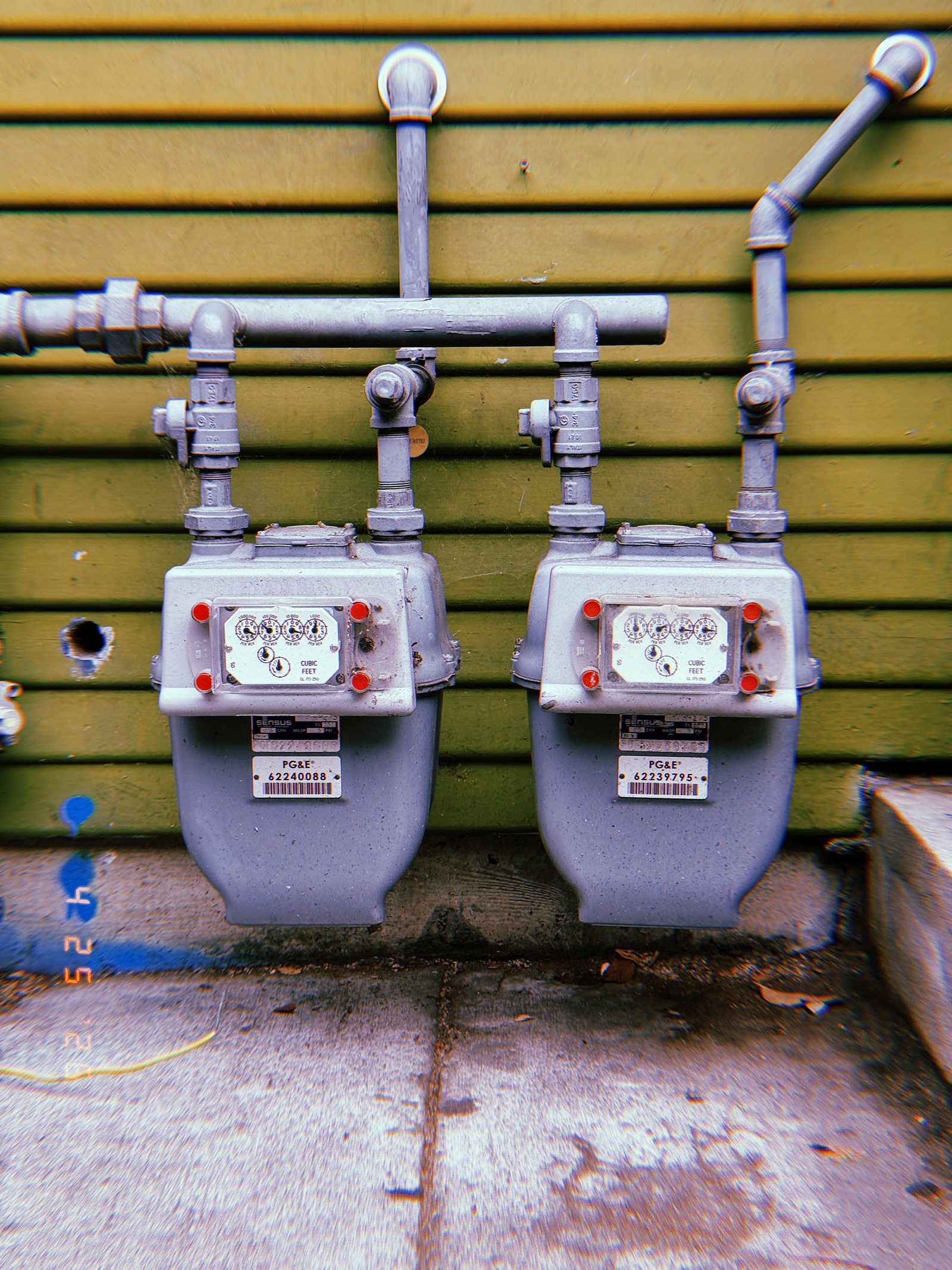 Two residential gas meters mounted side by side beneath exposed utility pipes on a green wooden exterior wall.