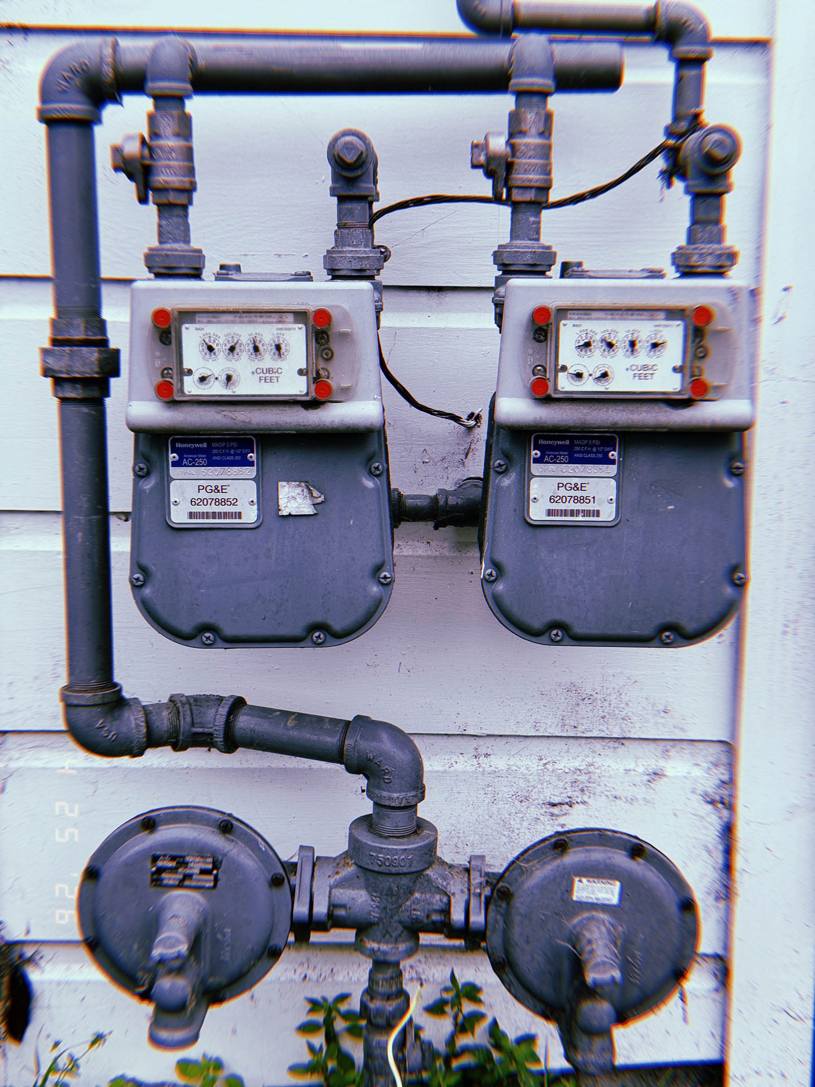 Two residential gas meters connected to an exposed pipe distribution system mounted on a white exterior wall above dual gas pressure regulators.
