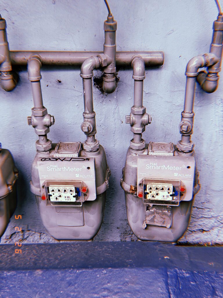 Two residential PG&E SmartMeter gas meters mounted against a light painted wall with exposed vertical pipe connections and valve infrastructure.