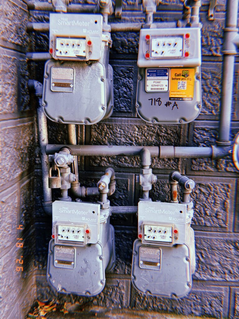 Four PG&E SmartMeter gas meters mounted on a dark textured exterior wall with interconnected pipe infrastructure, shutoff valves, and utility labeling.