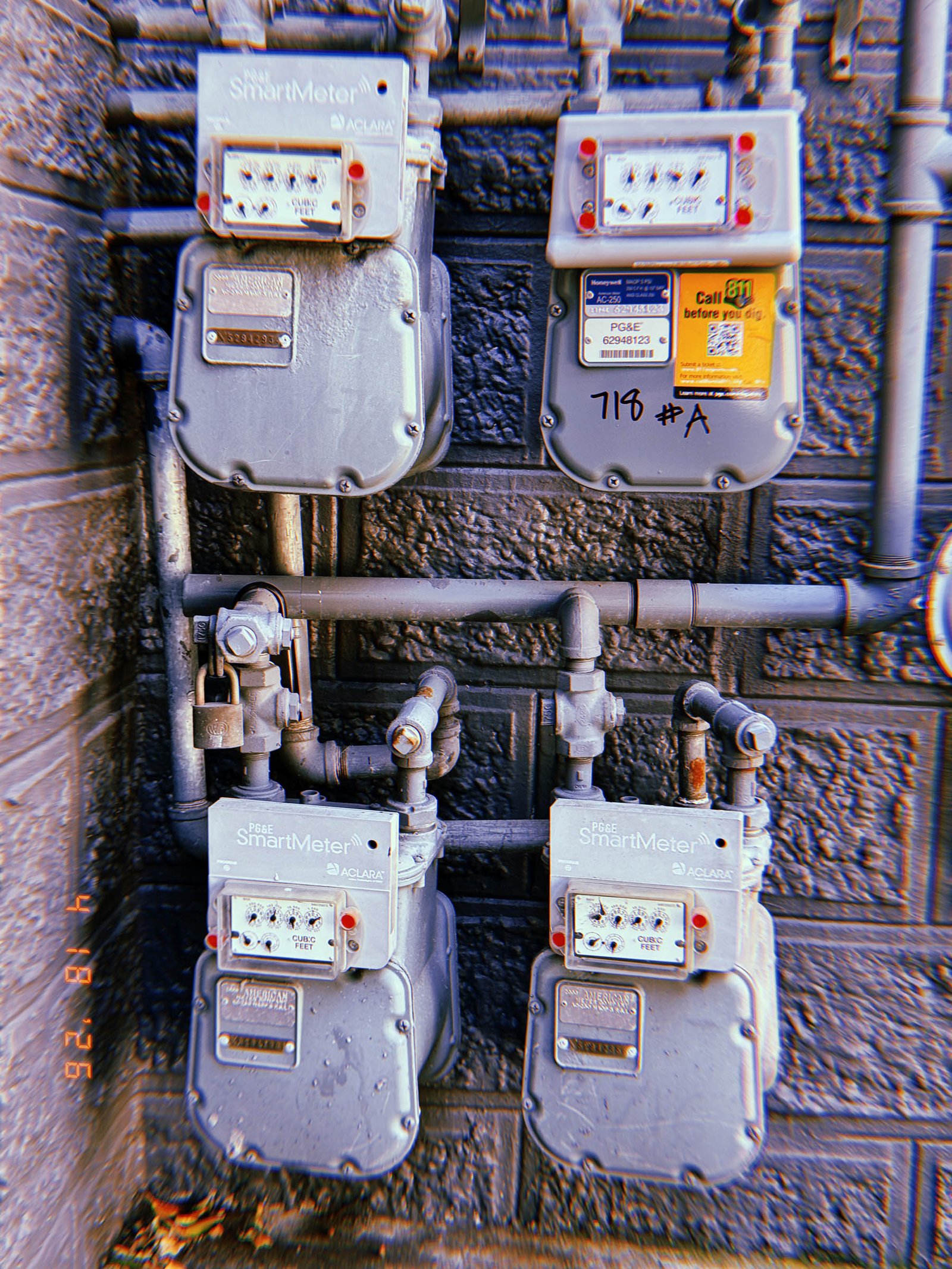 Four PG&E SmartMeter gas meters mounted on a dark textured exterior wall with interconnected pipe infrastructure, shutoff valves, and utility labeling.