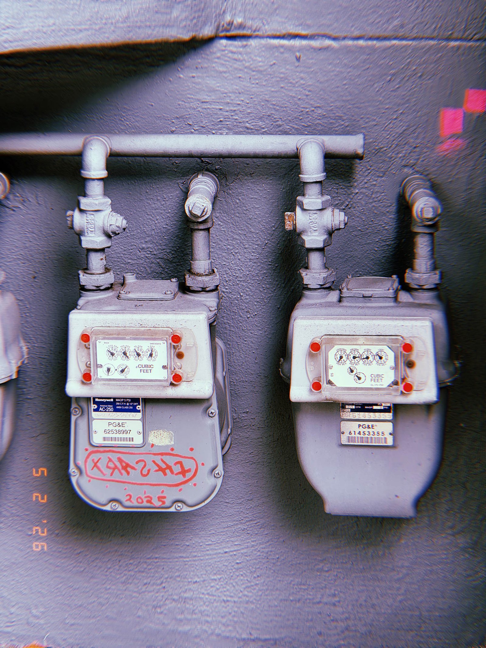 Two residential gas meters mounted on a dark painted wall with exposed pipe connections, shutoff valves, and red graffiti markings on one meter housing.