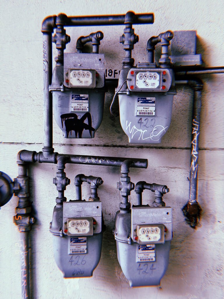 Four residential PG&E SmartMeter gas meters mounted on a concrete wall with exposed pipe connections, graffiti markings, and valve infrastructure.