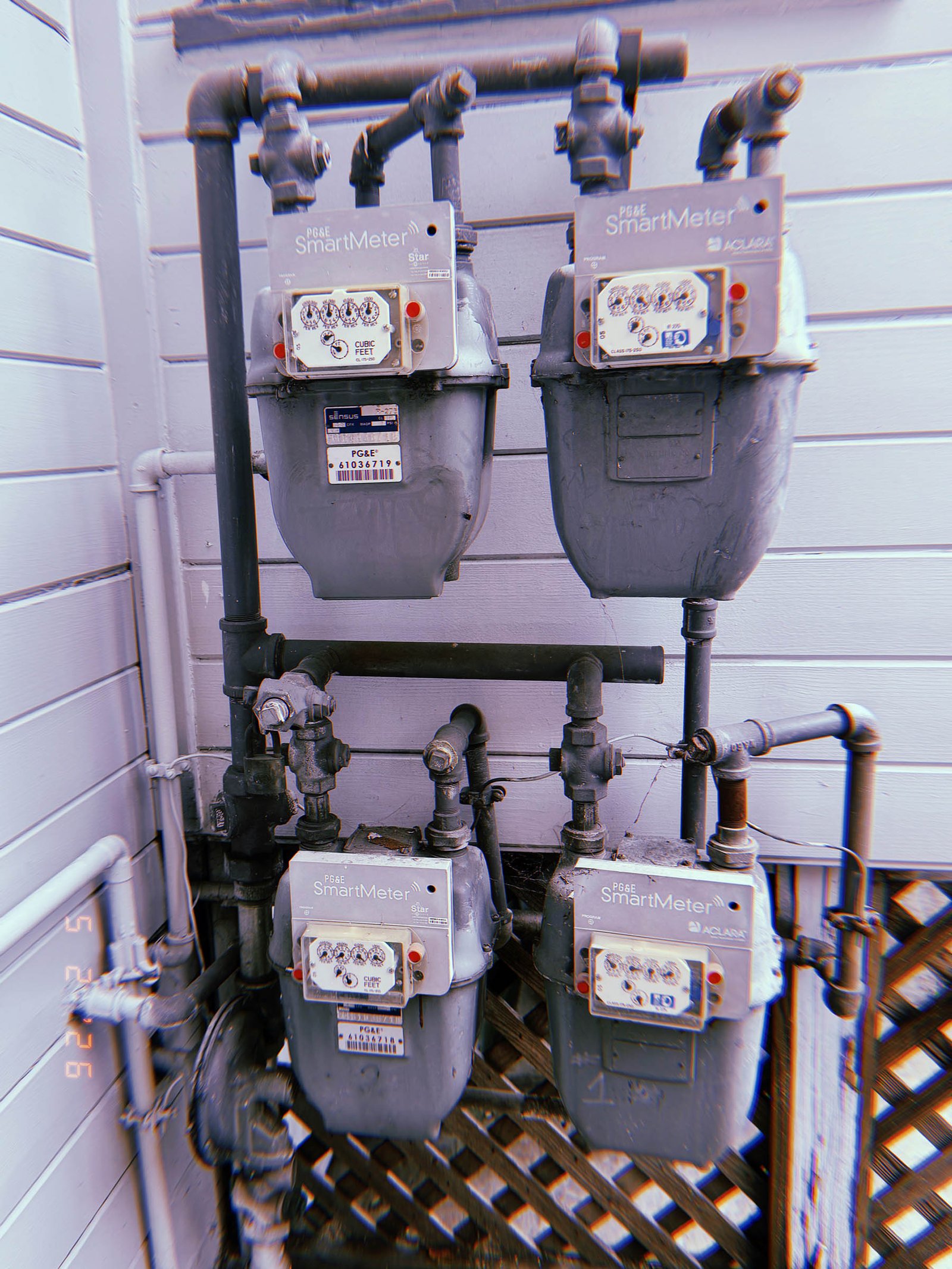 Four PG&E SmartMeter gas meters mounted on an exterior residential wall with interconnected black pipe infrastructure and valve assemblies.