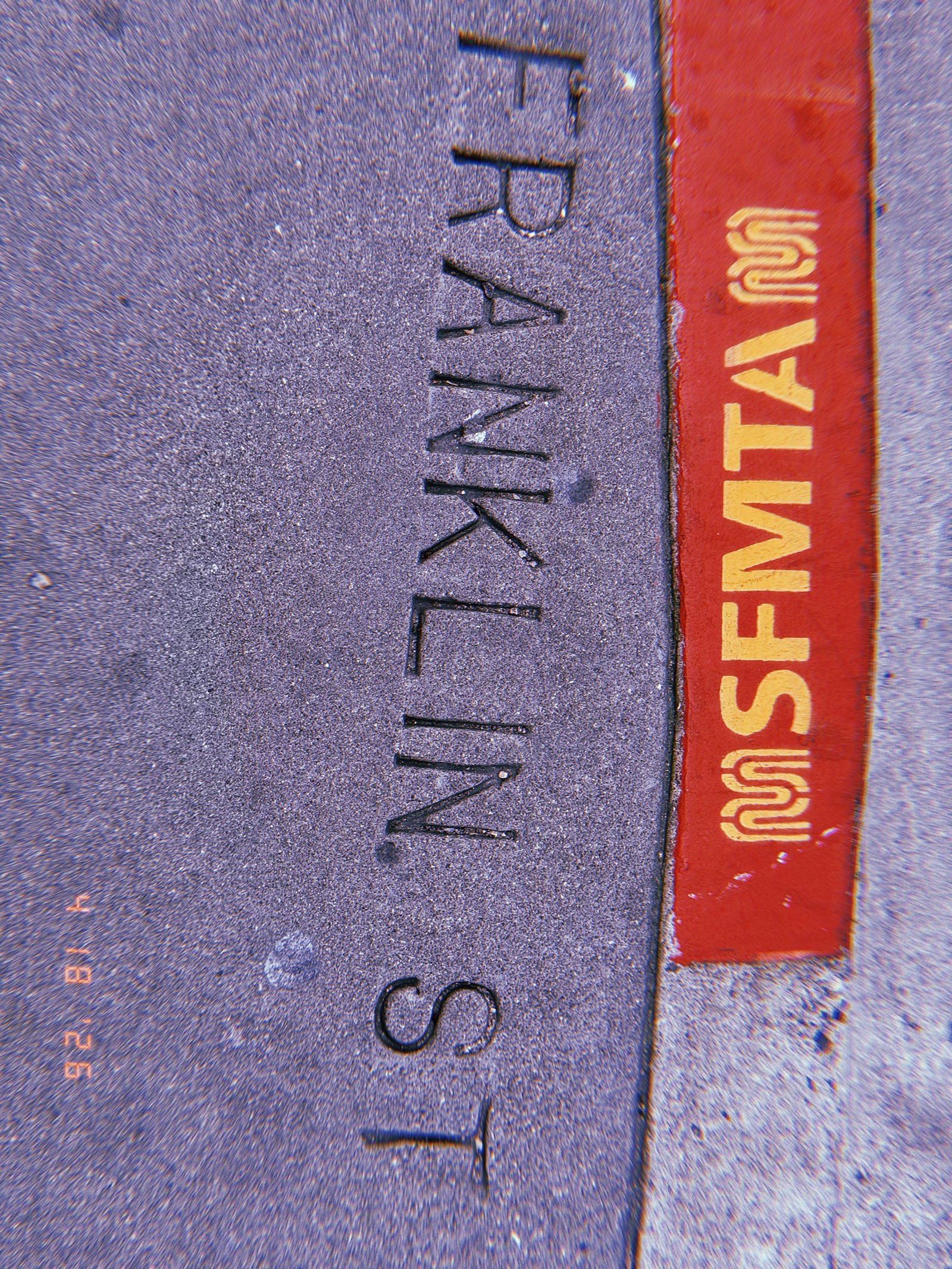 The words "FRANKLIN ST" embedded in a concrete sidewalk beside a red curb marked "SFMTA."