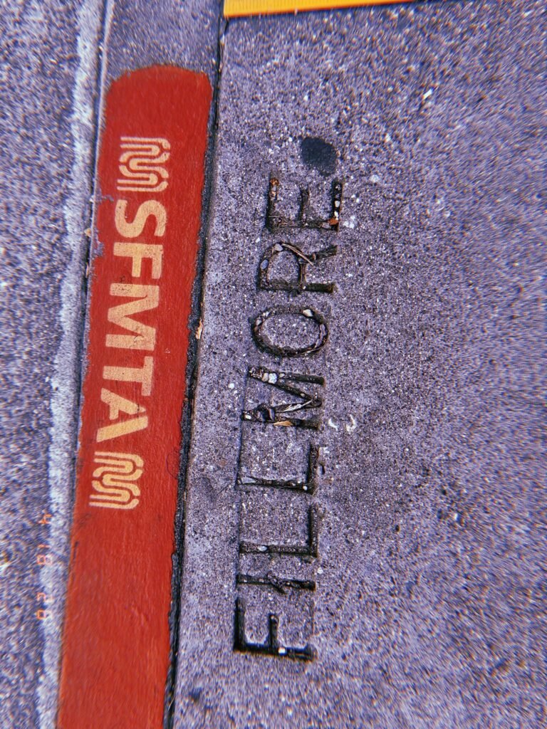 The word "FILLLMORE" embedded in a concrete sidewalk beside a red curb marked "SFMTA."