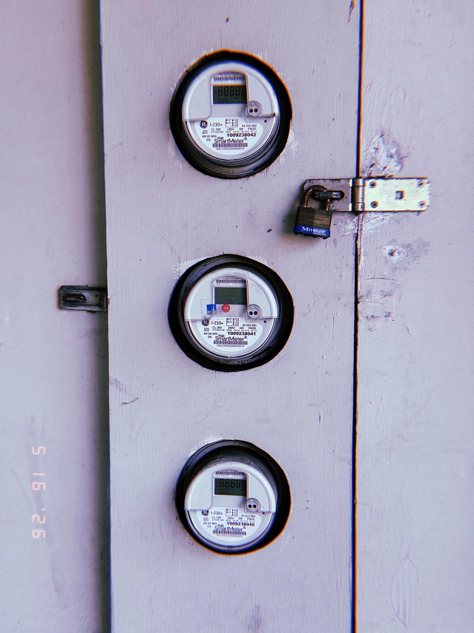 Three digital electrical smart meters embedded vertically within a painted utility enclosure door. A padlock secures the cabinet access panel, while the meters are arranged in a linear column against a flat industrial surface.